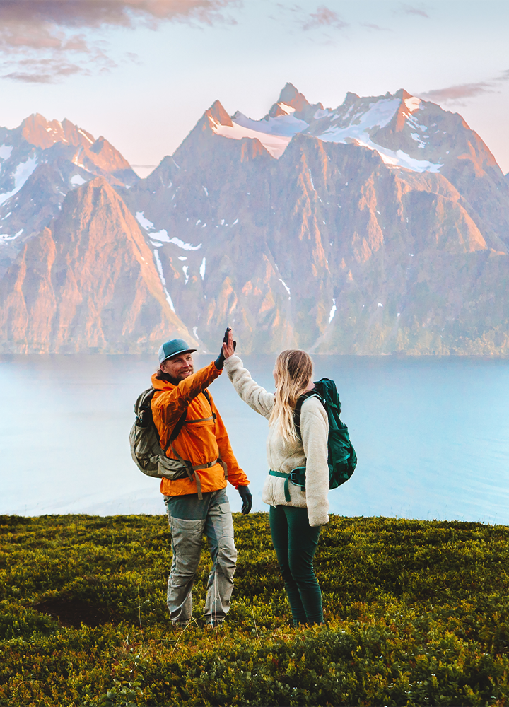 Dos excursionistas con mochilas se chocan las manos en frente a un lago rodeado de montañas nevadas
