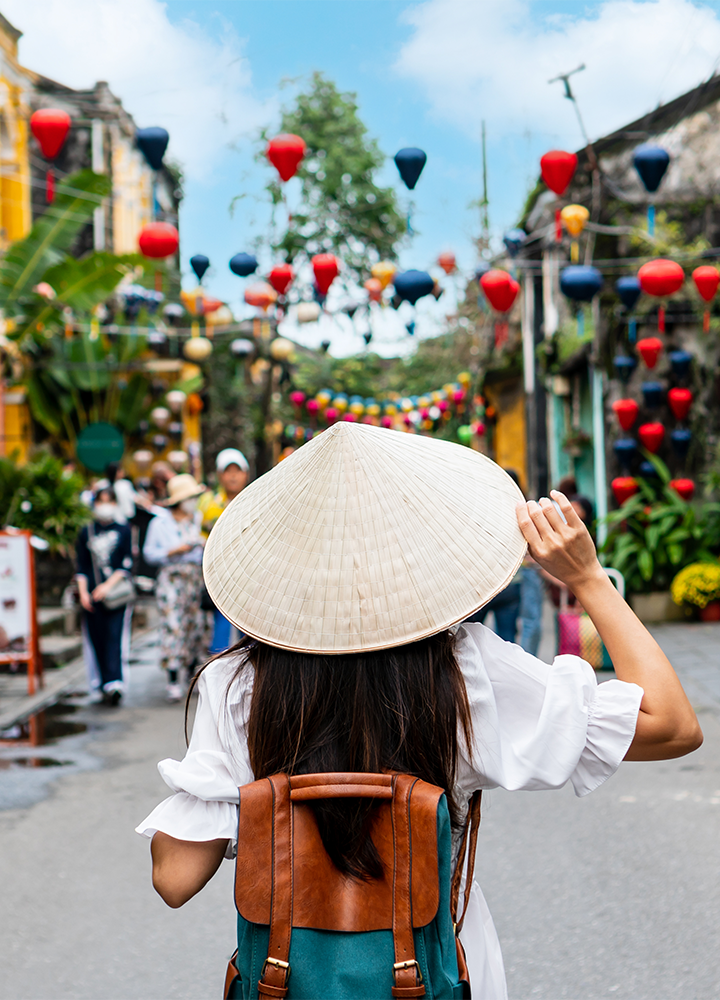 Persona con sombrero cónico vietnamita y mochila camina por una calle decorada con faroles rojos y coloridos.