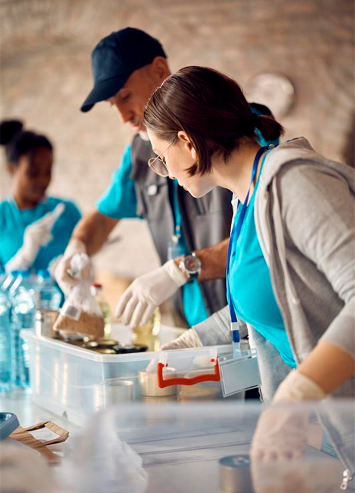 Varias personas con camisetas azules y guantes participan en un voluntariado, empacando agua, arroz y latas en cajas plásticas