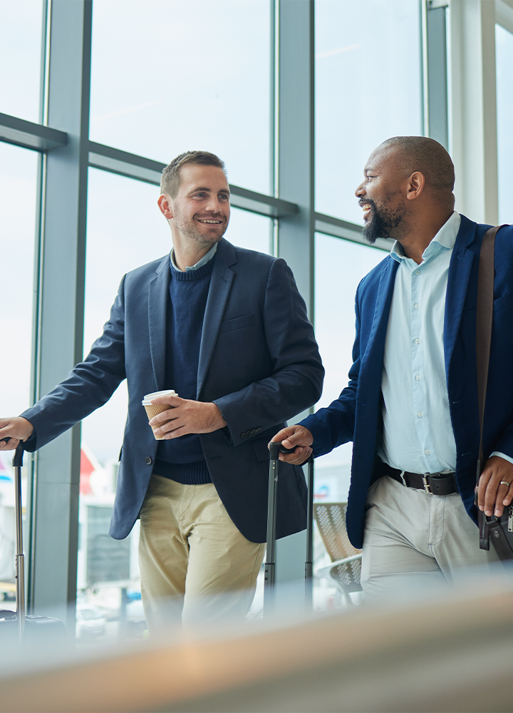 Dos hombres caminan por el aeropuerto con sus maletas; uno sostiene una taza de café.