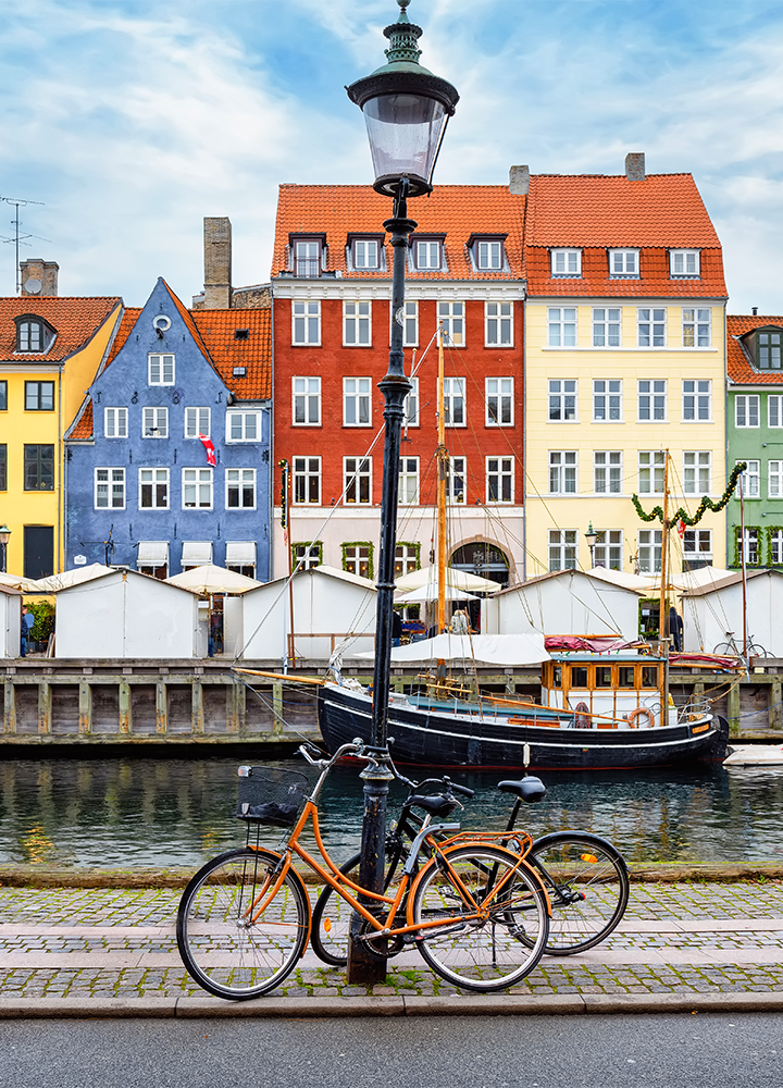 Calle en Copenhague junto a un canal, con bicicletas aparcadas, un poste de luz, edificios coloridos y un barco en el agua