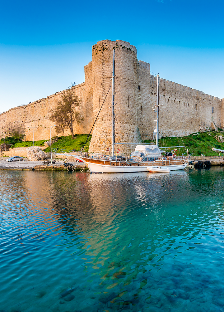 Castillo de piedra junto a un puerto tranquilo, donde hay un barco de vela atracado