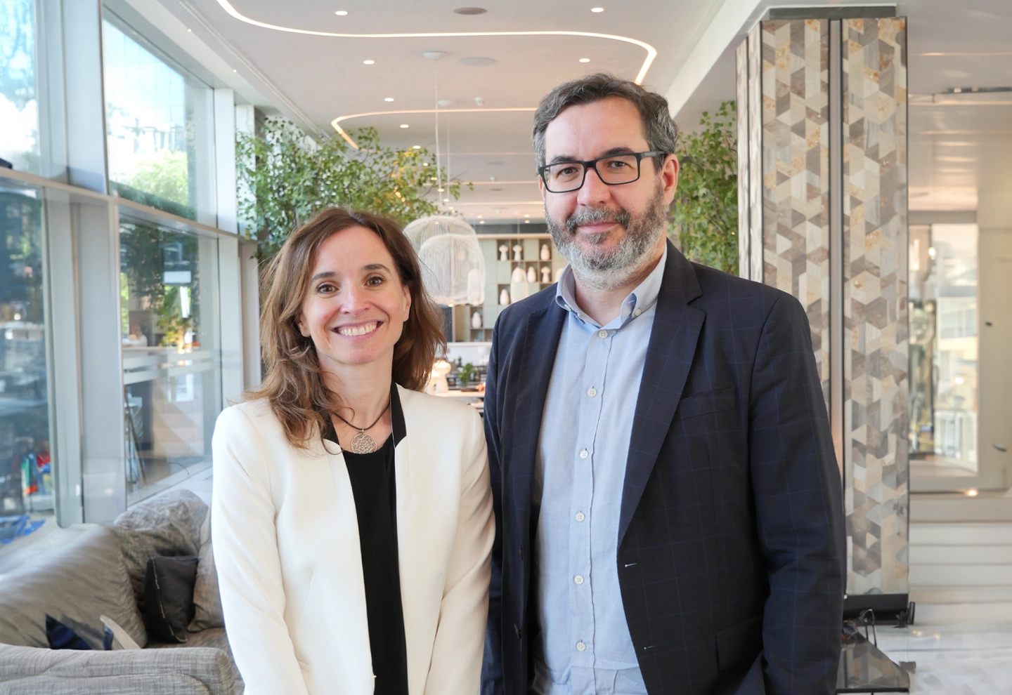 Two people stand inside a bright, modern room with large windows, plants, and stylish furniture—one wears a white blazer, the other a dark suit jacket