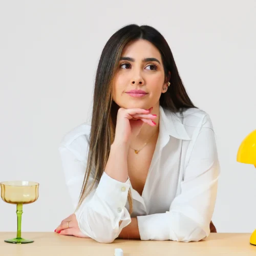 Una mujer con camisa blanca está sentada en una mesa con copas de base verde y parte superior amarilla; su mano toca el mentón con gesto reflexivo. A la derecha hay una lámpara amarilla de forma semiesférica, sobre fondo claro.
