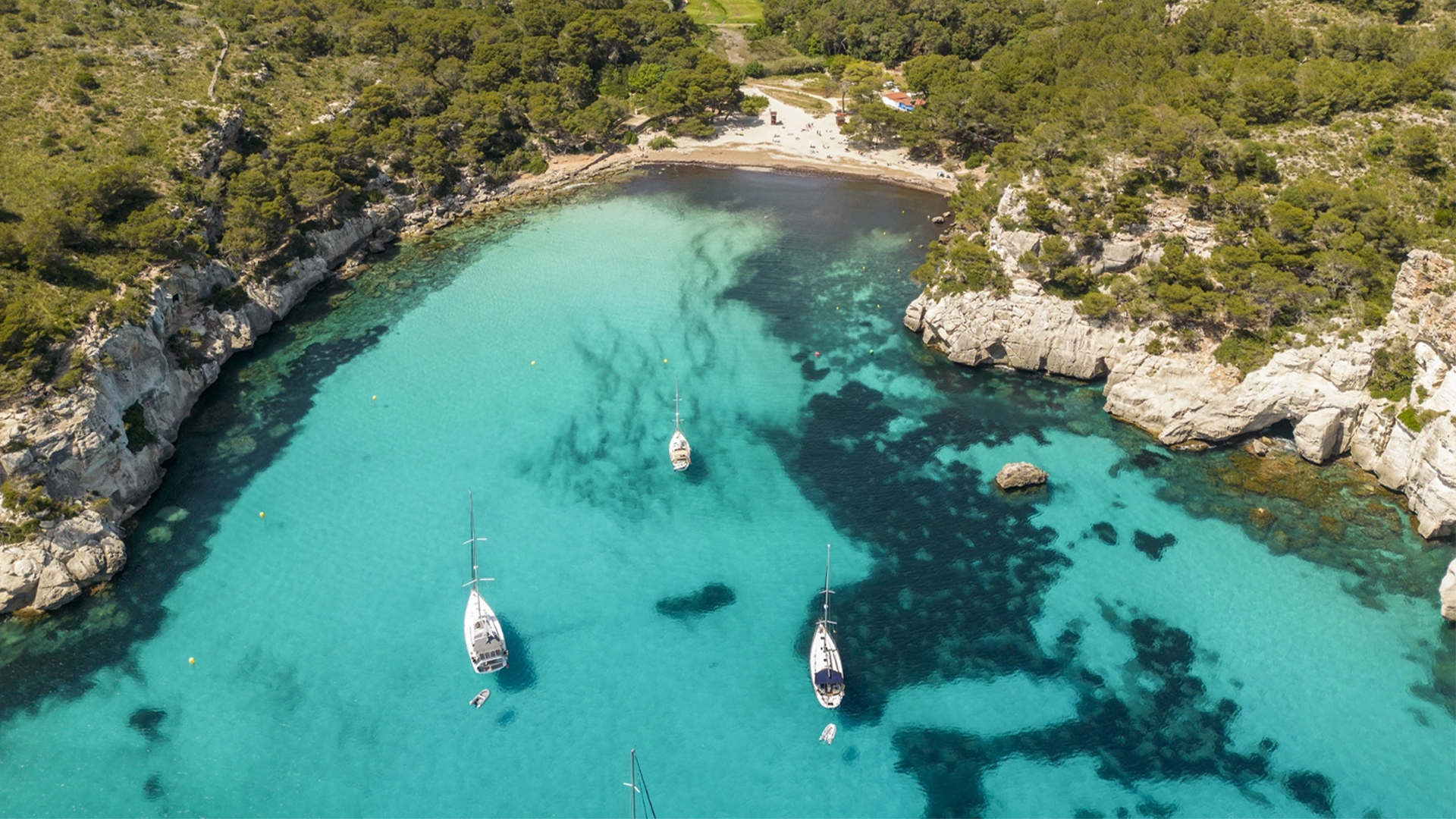 Vista aérea de una cala con aguas cristalinas, donde varios veleros blancos están anclados.