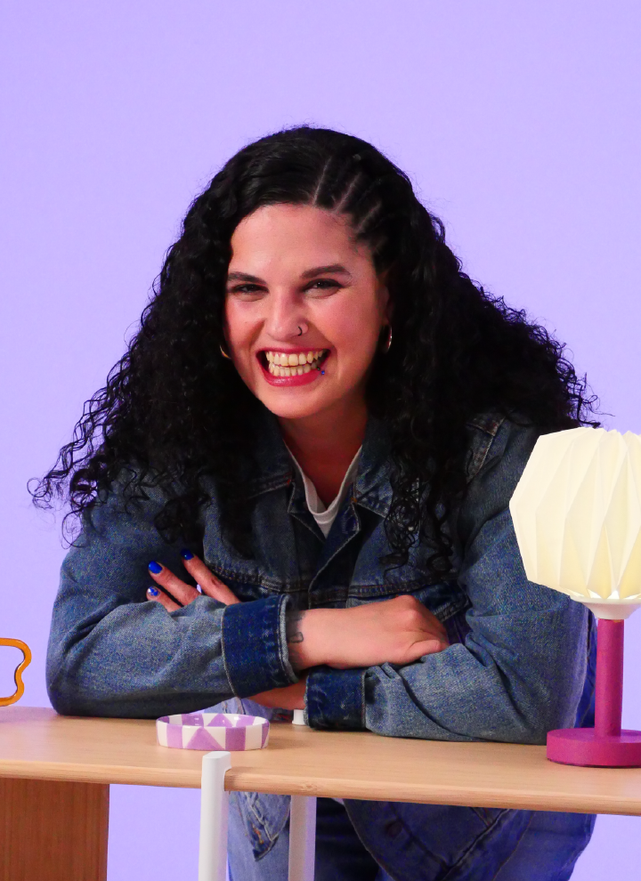 Smiling woman with long, curly hair, wearing a blue denim jacket and a white T-shirt. She is leaning on a table with her arms crossed, giving her a relaxed posture. On the table, there are several interesting objects: on the left, a small, irregularly shaped orange item; in the center, a circular object with a geometric pattern in purple and white tones; and on the right, a table lamp with a purple base and a white pleated lampshade. The background is a light lilac tone, creating a calm and harmonious atmosphere.
