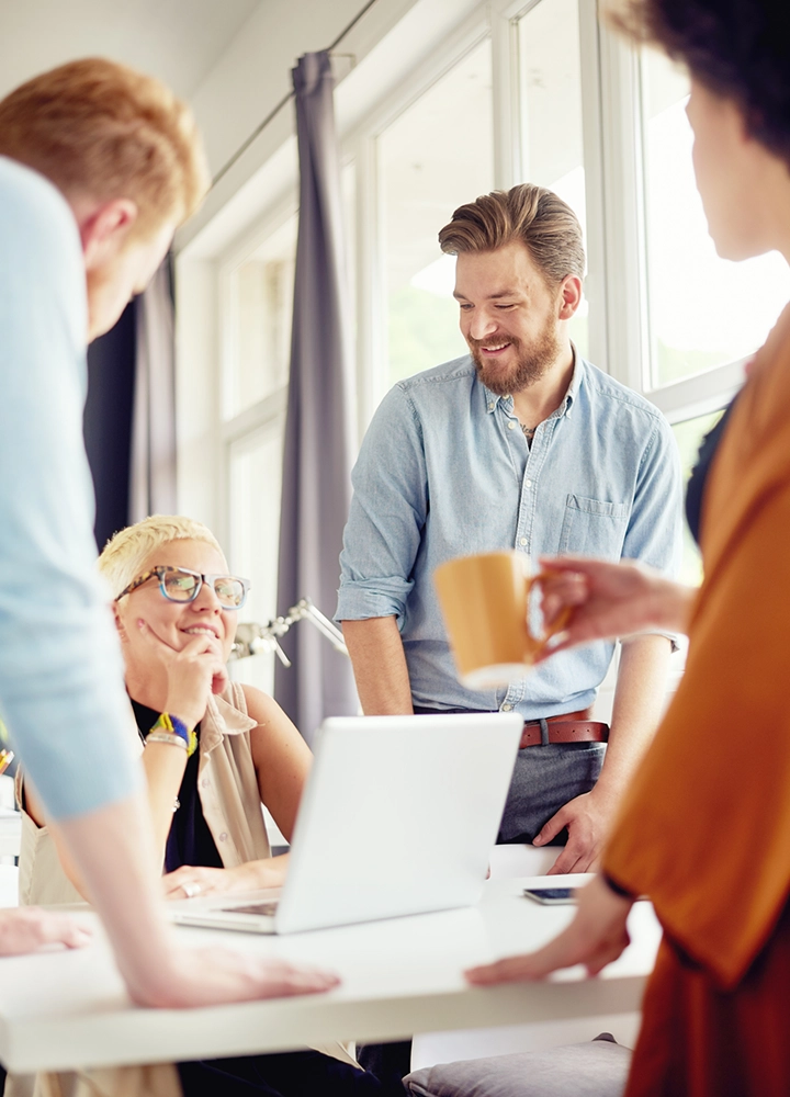 Creative work team: a woman with a laptop and a bearded man smiling in front of other colleagues.