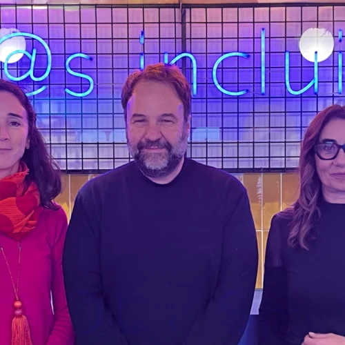 Three people pose together in front of a mesh backdrop and a blue neon sign that reads “Tod@s incluíd@s” (All included).