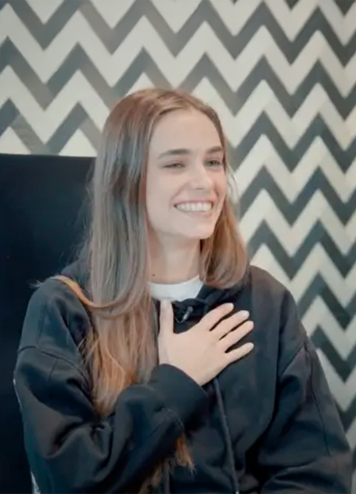 A person with long hair, wearing a white T-shirt and a black sweatshirt, sits in front of a black-and-white zigzag background, with one hand resting on their chest.
