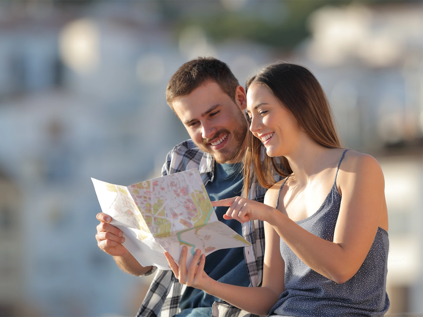 Una pareja sonriente está sentada al aire libre mirando un mapa de papel juntos. La mujer señala un punto del mapa mientras el hombre lo sostiene.