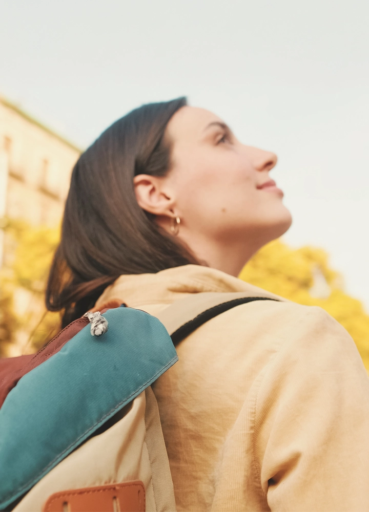 Una mujer joven, vista de perfil, mira hacia arriba con una expresión de asombro. Lleva una mochila con detalles en marrón y azul y un jersey beige. El sol brilla desde un lado.
