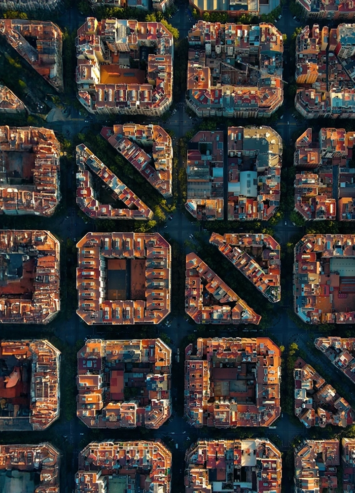 Vista aérea de Barcelona con sus característicos bloques de edificios cuadriculados y patios interiores. Se ven las azoteas, las calles y la vegetación de los patios.