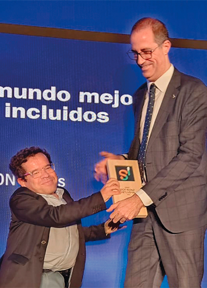 Two people are on a stage; one is handing an award to the other, while a screen behind them reads: “Building a better world with everyone included” and “From ILUNION.”