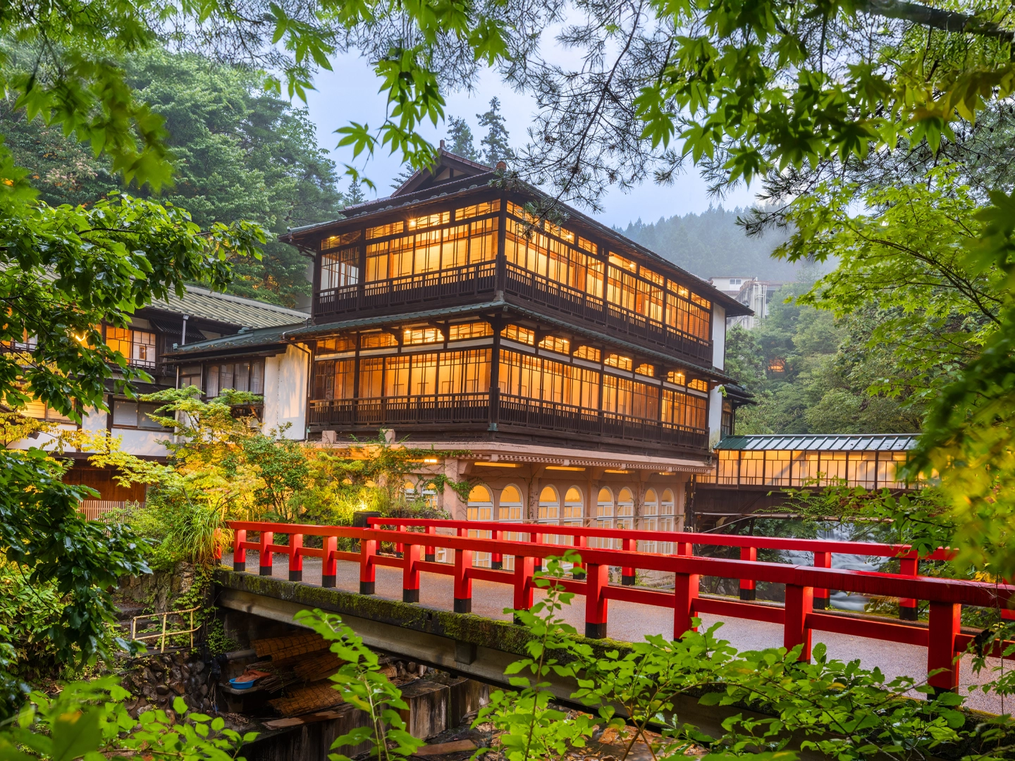 Un ryokan tradicional japonés de madera, con las luces interiores encendidas, detrás de un puente rojo que cruza un arroyo.