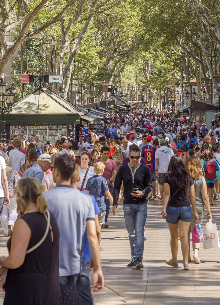 Una vista concurrida de La Rambla, en Barcelona. Hay una multitud de gente caminando por el paseo central, con árboles frondosos y quioscos a ambos lados.