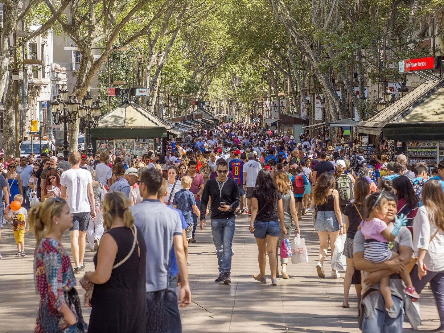Una vista concurrida de La Rambla, en Barcelona. Hay una multitud de gente caminando por el paseo central, con árboles frondosos y quioscos a ambos lados.