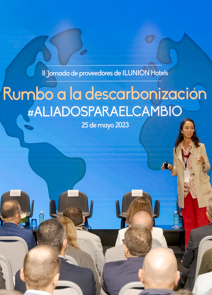 Sala con personas sentadas escucha a mujer con micrófono frente a una pantalla grande que dice: “II Jornada de proveedores de ILUNION Hotels. Rumbo a la descarbonización. #ALIADOSPARAELCAMBIO. 25 de mayo 2023”.
