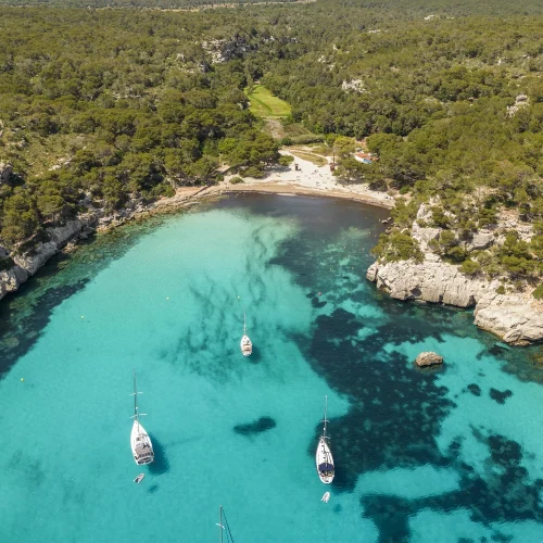 A panoramic aerial view of a sandy cove surrounded by cliffs and forests, with bright turquoise water. Several sailboats are floating in the calm bay.