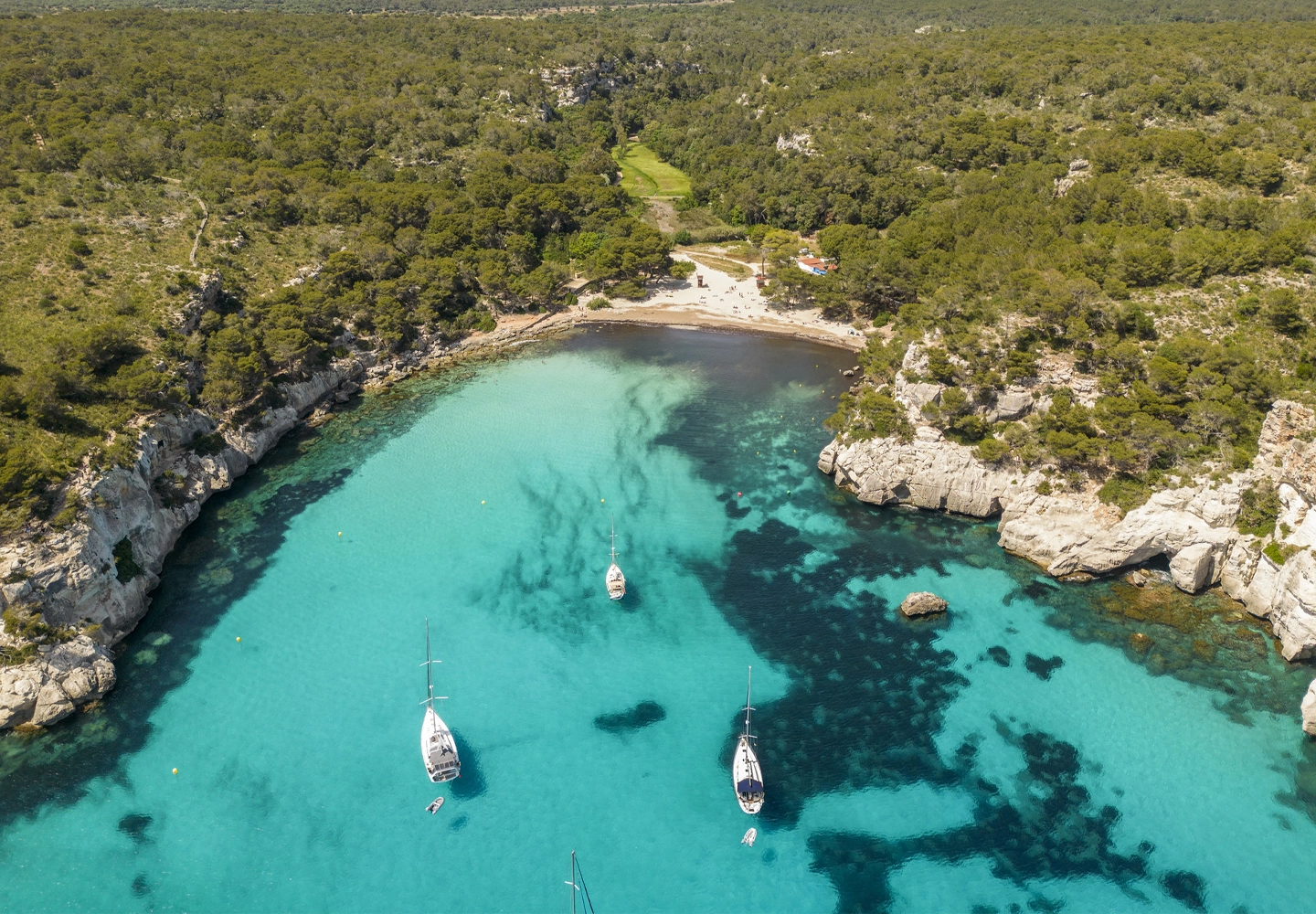 A panoramic aerial view of a sandy cove surrounded by cliffs and forests, with bright turquoise water. Several sailboats are floating in the calm bay.