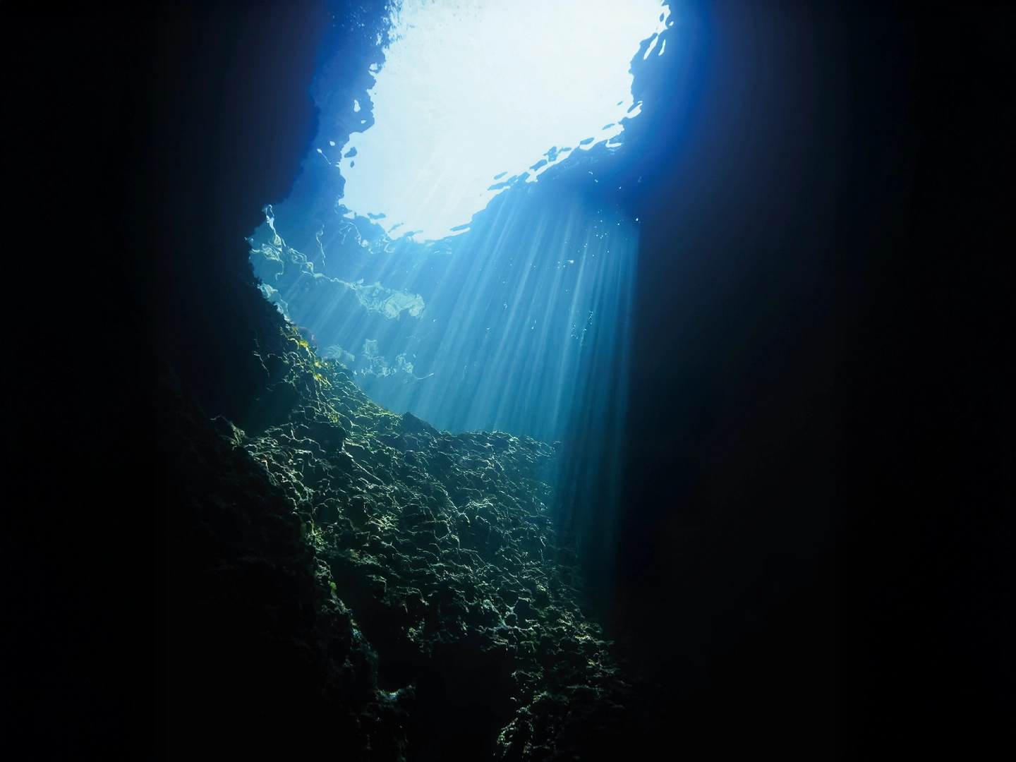 Luz solar entra desde la superficie a través de un agujero en una cueva submarina. Los rayos de luz iluminan el agua azul y los corales o rocas de la pared de la cueva.