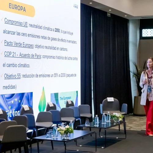 Sala de conferencias con sillas frente a una pantalla que muestra compromisos climáticos de la Unión Europea, mientras una mujer está de pie en el escenario hablando al público.