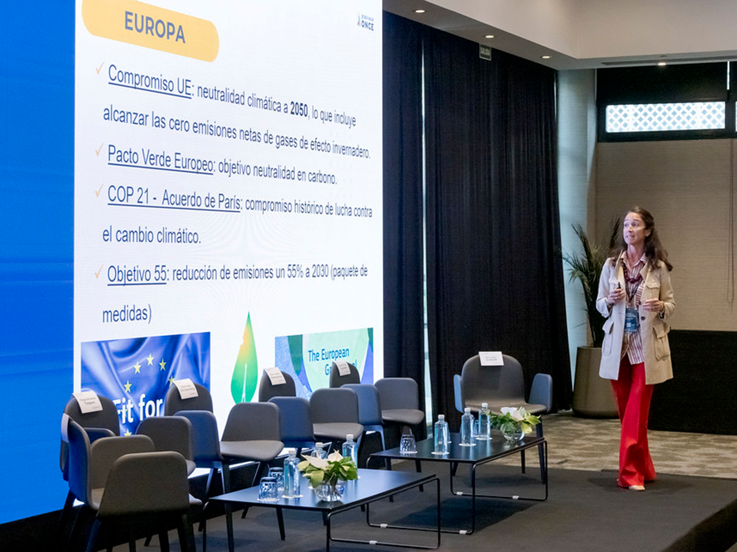 Sala de conferencias con sillas frente a una pantalla que muestra compromisos climáticos de la Unión Europea, mientras una mujer está de pie en el escenario hablando al público.