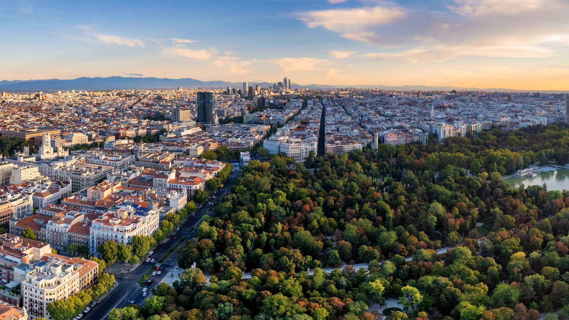 Imagen muestra una vista aérea de Madrid al atardecer, con la ciudad extendiéndose en el fondo repleta de edificios altos, y junto a ella el Parque del Retiro, un espacio verde con árboles frondosos.