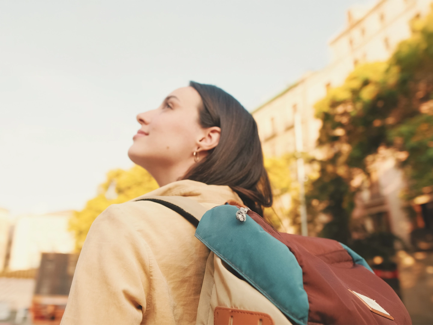Una mujer joven, vista de perfil, mira hacia arriba con una expresión de asombro. Lleva una mochila con detalles en marrón y azul y un jersey beige. El sol brilla desde un lado.