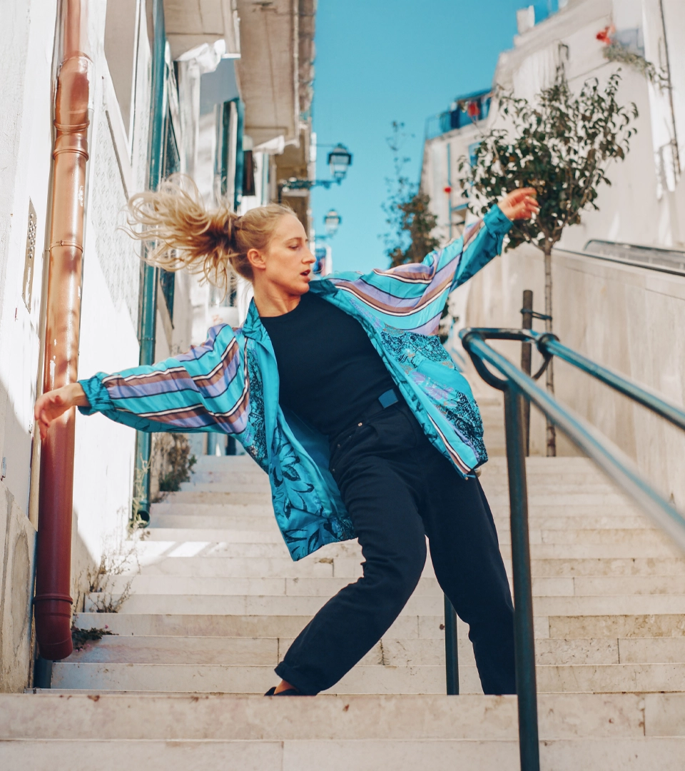 A smiling blonde woman, dressed in a blue silk jacket and black pants, gracefully dances down an outdoor stone staircase.