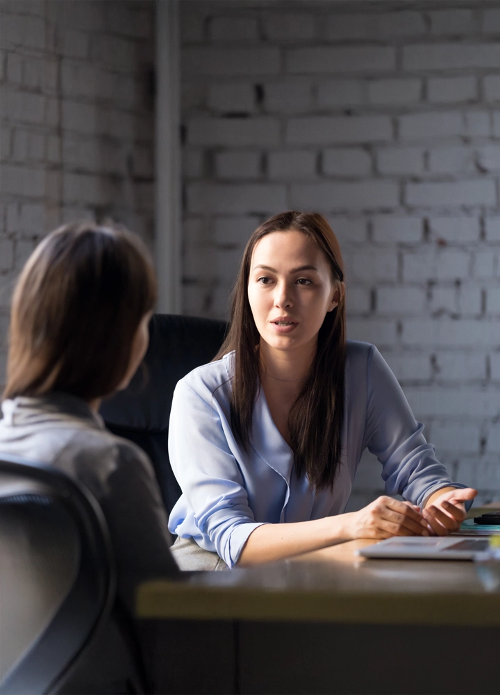 Dos mujeres están sentadas frente a frente en un escritorio, con una de ellas hablando y gesticulando. El fondo muestra una pared de ladrillos blancos.