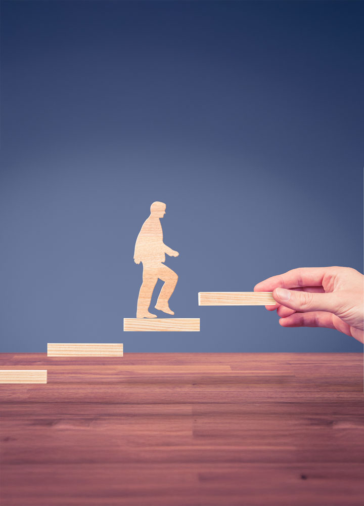 Una silueta de madera de una persona sube una escalera de madera. Una mano ayuda a la silueta colocando la última pieza del escalón. El fondo es azul oscuro.