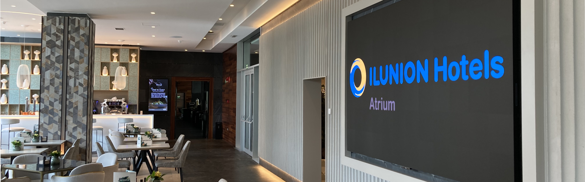 Sala moderna y luminosa con mesas, plantas pequeñas, lámparas colgantes y una pantalla que muestra el texto “ILUNION Hotels Atrium”.