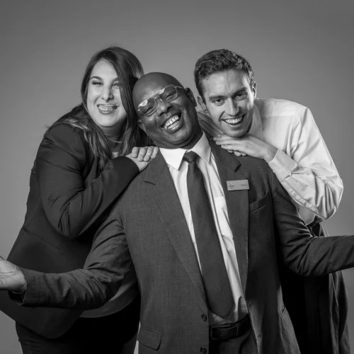 Three elegantly dressed people pose together; the one in the center wears a suit and tie with arms outstretched, while the other two rest a hand on each of his shoulders. The image is in black and white.