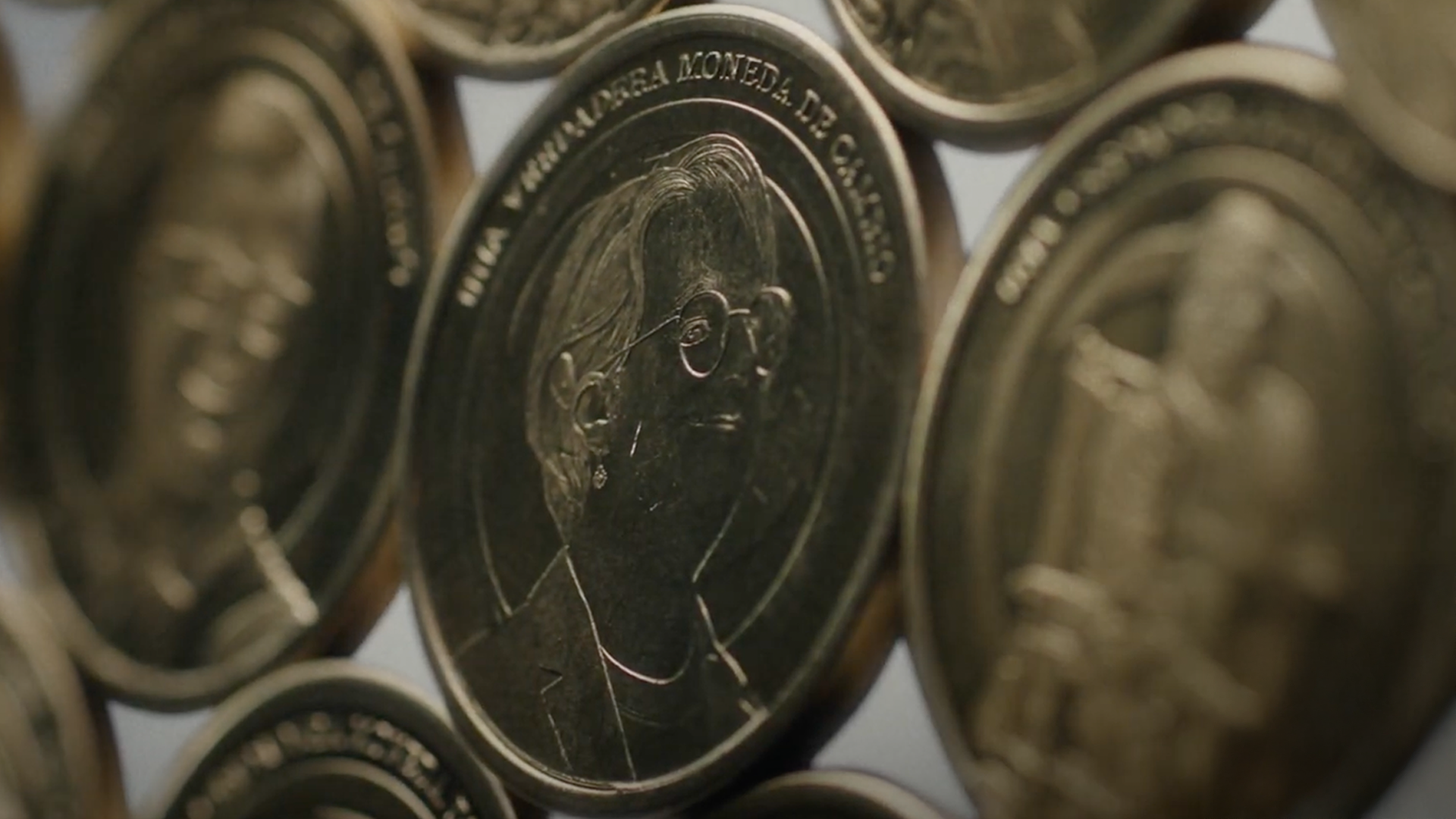 Primer plano de monedas con perfil grabado de persona con gafas. Texto: “UNA GRANDE MONEDA DE CAMBIO.” Diseño detallado y metálico.