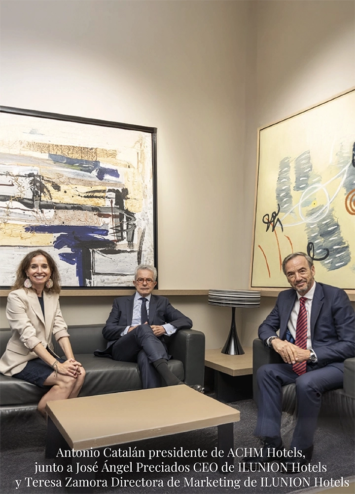 Tres personas en reunión formal, sentadas en sillones negros alrededor de mesa baja. Fondo con arte abstracto y lámpara moderna. Ambiente profesional.