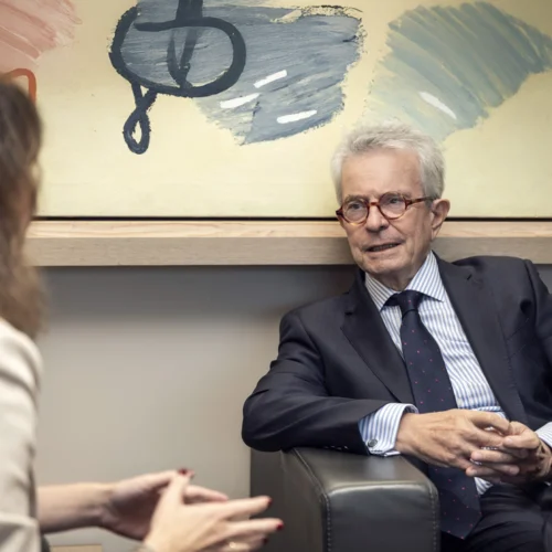 Un hombre mayor de pelo gris con gafas, traje y corbata se sienta en un sillón, mirando y hablando con una mujer cuyas manos se ven de espaldas. Detrás, una pintura abstracta.
