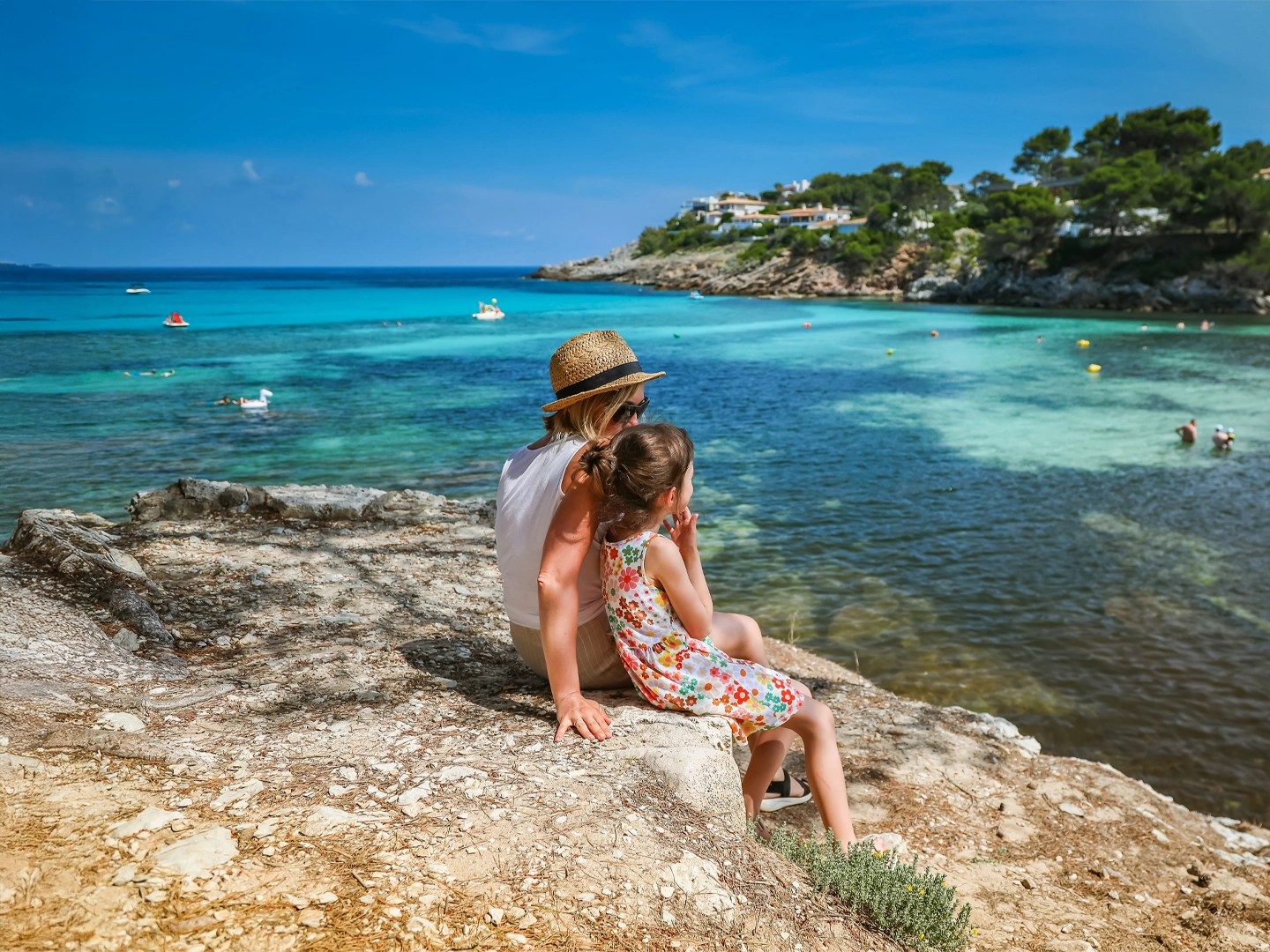 Una mujer con un sombrero de paja y una niña están sentadas en una roca, contemplando una cala de aguas turquesas en un día soleado. Hay barcos y casas en la orilla.