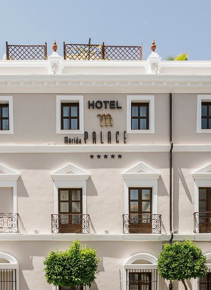 La imagen muestra la fachada del HOTEL Mérida PALACE, con arquitectura clásica y varias ventanas con balcones decorativos. Bajo el nombre aparecen cinco estrellas. En la parte superior hay una terraza con plantas, y al nivel de la calle se ven árboles.