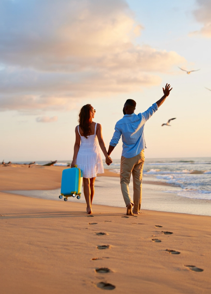 Dos personas caminando de la mano por la playa al atardecer, una con vestido blanco y maleta azul, otra con camisa celeste y brazo levantado.
