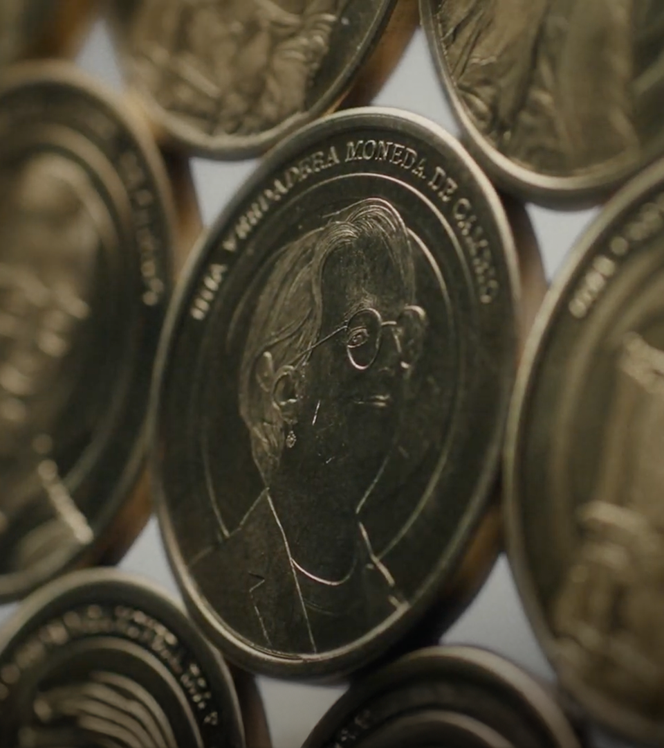 Primer plano de monedas con perfil grabado de persona con gafas. Texto: “UNA GRANDE MONEDA DE CAMBIO.” Diseño detallado y metálico.