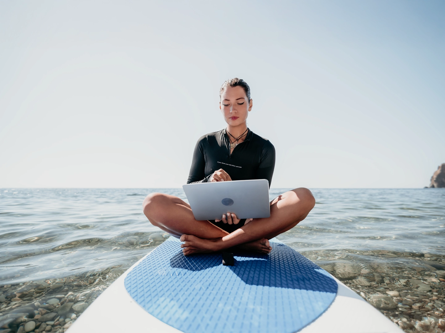 Persona sentada de frente en una tabla de paddle surf sobre agua tranquila, usando portátil. Día soleado, entorno natural y sereno que refleja trabajo remoto relajado.