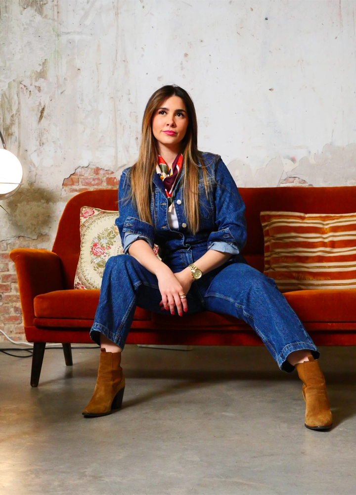 Plano completo de una mujer sentada en un sofá naranja, vistiendo un mono de mezclilla, botines de gamuza marrón y una pañoleta, con una pared de ladrillo desgastado detrás.