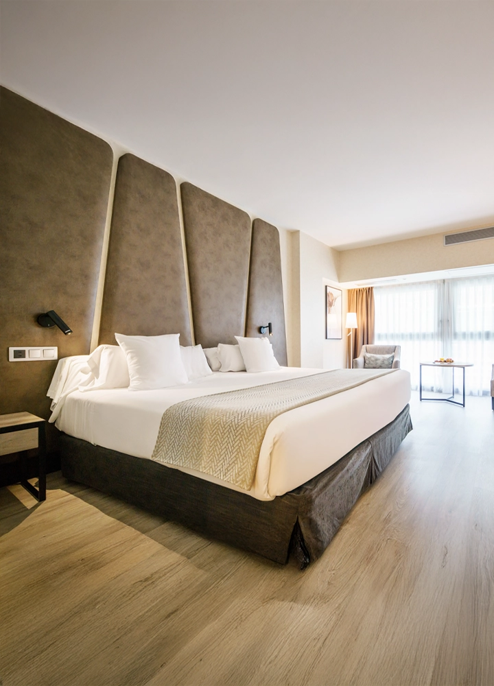 Una habitación de hotel moderna y espaciosa. Tiene una cama grande con un cabecero acolchado, dos sillones, un escritorio, una gran ventana con cortinas y suelo de madera.