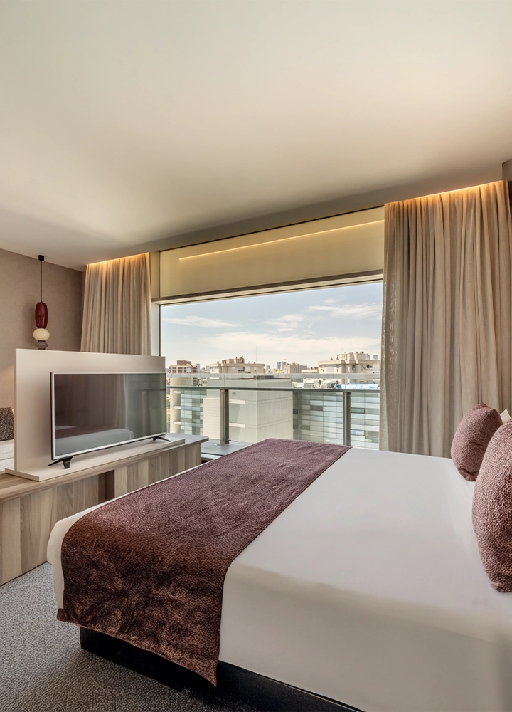 Una habitación de hotel moderna con una cama grande y un televisor de pantalla plana. Hay grandes ventanales con vistas a un paisaje urbano, y la cama tiene una manta de color marrón.