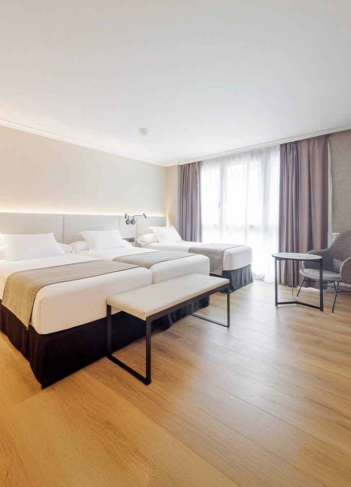 Una habitación de hotel luminosa con tres camas individuales y un banco de pie de cama. Hay grandes ventanas con cortinas marrones y el suelo es de madera.