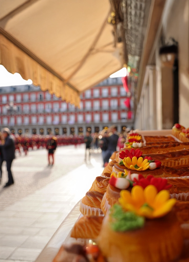 Primer plano de pasteles decorados con flores coloridas. Están sobre una mesa en una plaza con gente y edificios rojos y blancos al fondo. Ambiente festivo.