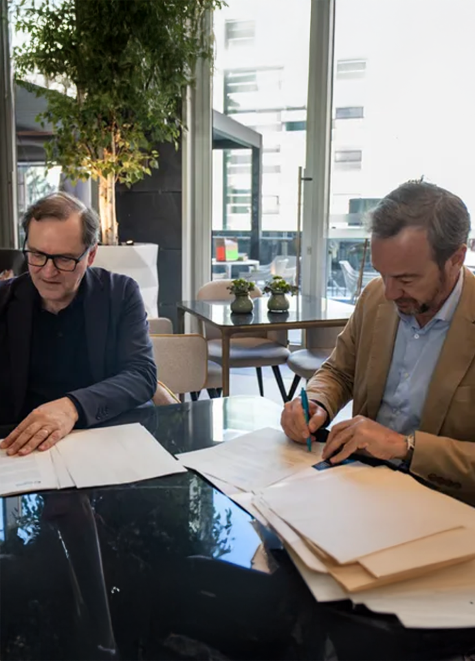 Two men seated at a table reviewing and signing documents in a professional setting with natural light and decorative plants.