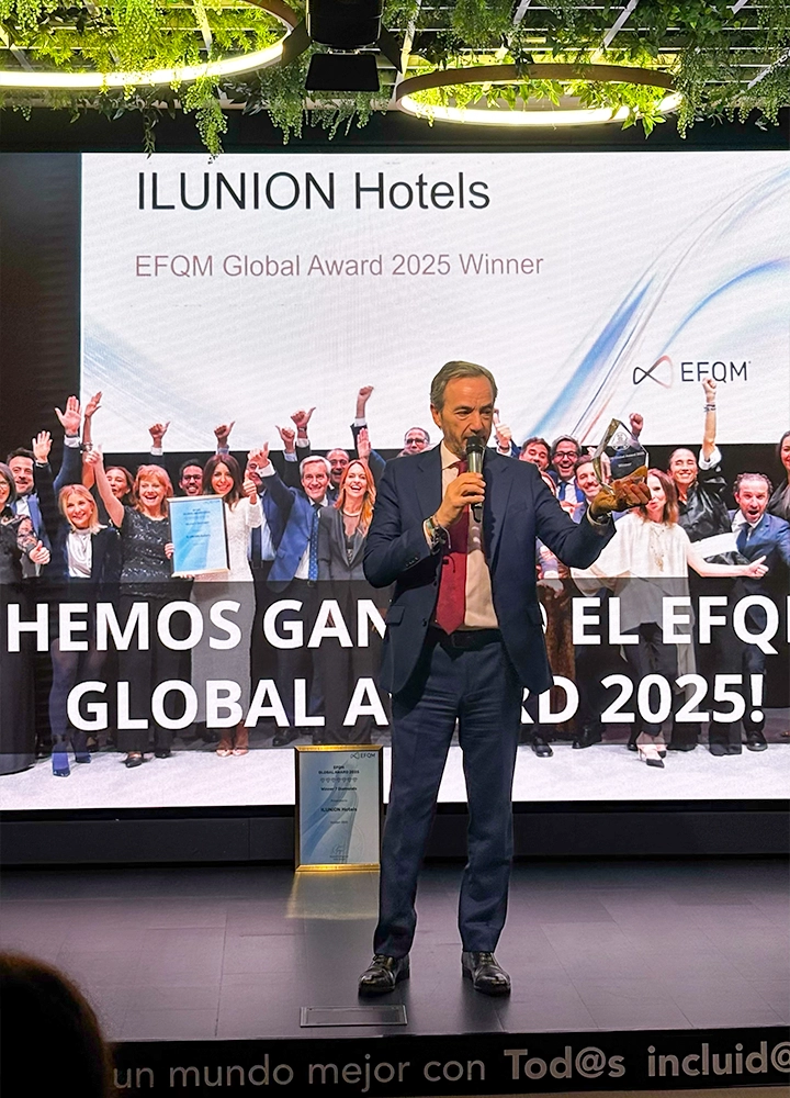 Un hombre en un escenario sosteniendo un trofeo frente a pantalla que anuncia: ILUNION Hotels, ganador del EFQM Global Award 2025, con grupo celebrando detrás.