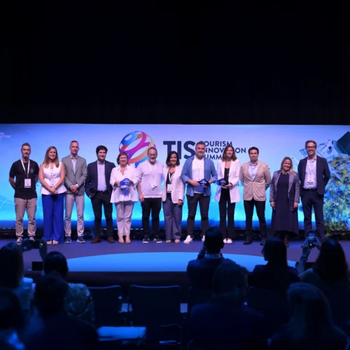 Grupo de personas en el escenario del Tourism Innovation Summit, posando frente a un fondo azul con el logo del evento y público en primer plano.