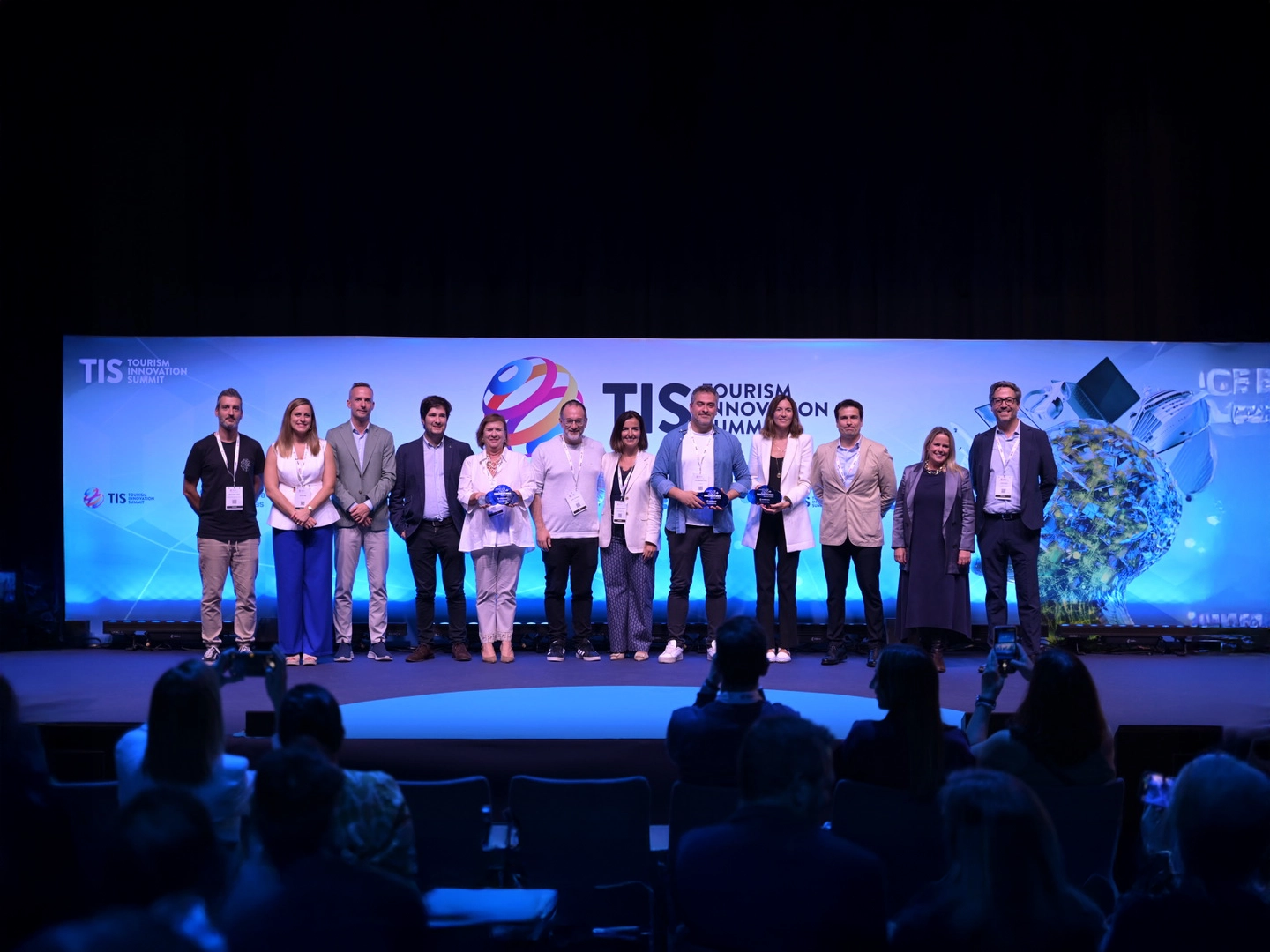 Grupo de personas en el escenario del Tourism Innovation Summit, posando frente a un fondo azul con el logo del evento y público en primer plano.